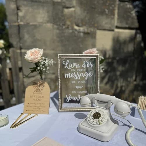 Livre d’or audio : téléphone rétro blanc posé sur une table de mariage, à côté d’un cadre « Livre d’or » et de roses