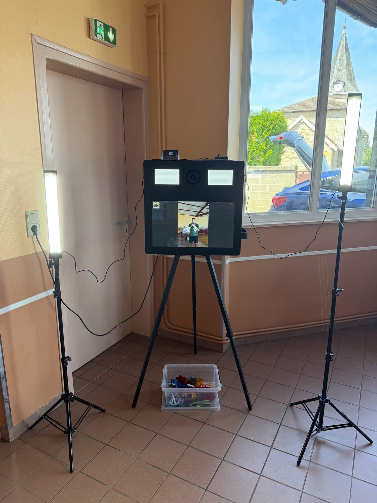 Photobooth classique noir sur trépied, entre deux éclairages LED, dans une salle des fêtes, avec sa caisse d'accessoires