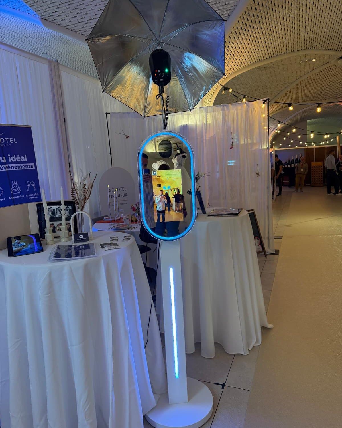 Photobooth miroir compact installé sur un salon : miroir ovale au contour lumineux bleu, colonne blanche